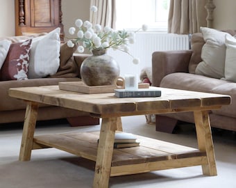 May include: A rustic, rectangular wooden coffee table with a lower shelf. The table is made of light-colored wood and has a natural finish. A vase with white flowers and a tray with decorative items sit on top. The table is in a living room setting.