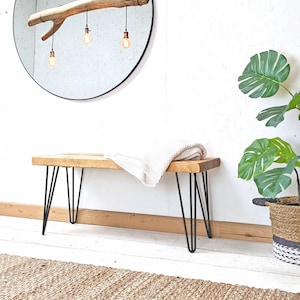 Op de afbeelding: Een houten bank met zwarte haarspeldpoten. De bank is gemaakt van gerecycled hout en heeft een natuurlijke afwerking. Een witte deken is over de bank gelegd. De bank staat voor een witte muur met een spiegel en een plant in een pot.