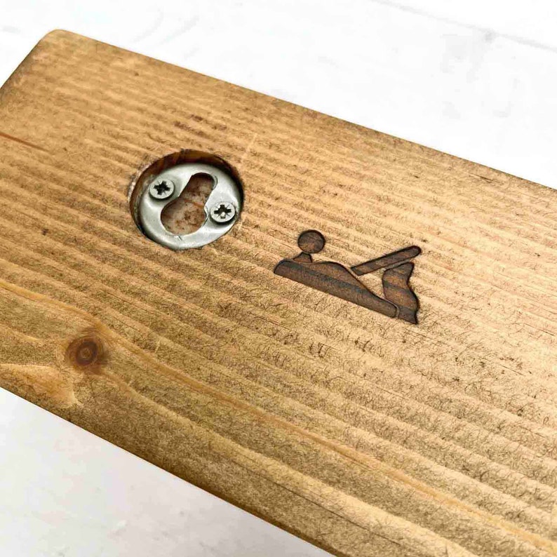 Op de afbeelding: Een houten plank met een flesopener en een klein, gegraveerd beeld van een handvlak. De plank is in een donkerbruine kleur gebeitst.