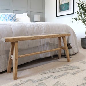 May include: A rustic wooden bench with a natural finish. The bench has a simple design with two legs on each side and a single support beam underneath. The bench is sitting on a light-colored rug in a bedroom setting.