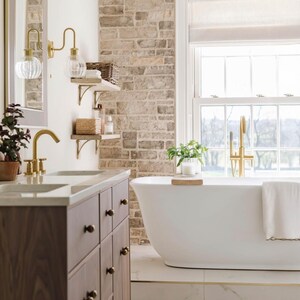 May include: A bathroom featuring a white bathtub, a wooden vanity, and a brick wall. The bathtub has a gold tap and a white towel. The vanity has a marble countertop and dark brown cabinets. Gold wall lights with glass globes are mounted on the wall.