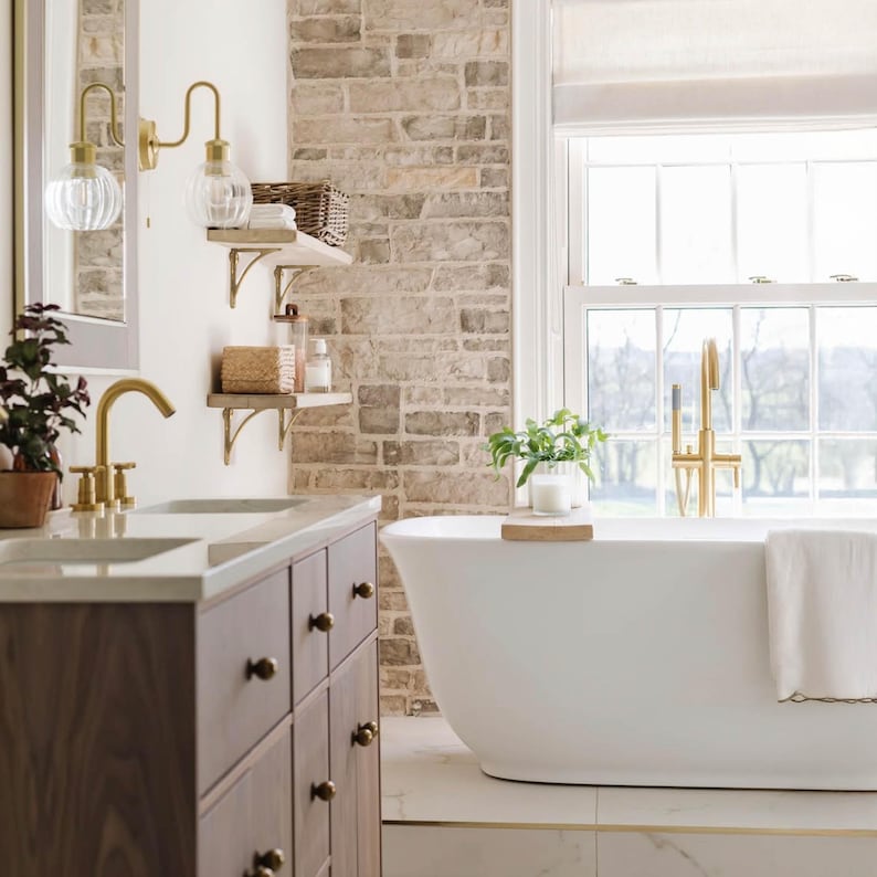 May include: A bathroom featuring a white bathtub, a wooden vanity, and a brick wall. The bathtub has a gold tap and a white towel. The vanity has a marble countertop and dark brown cabinets. Gold wall lights with glass globes are mounted on the wall.