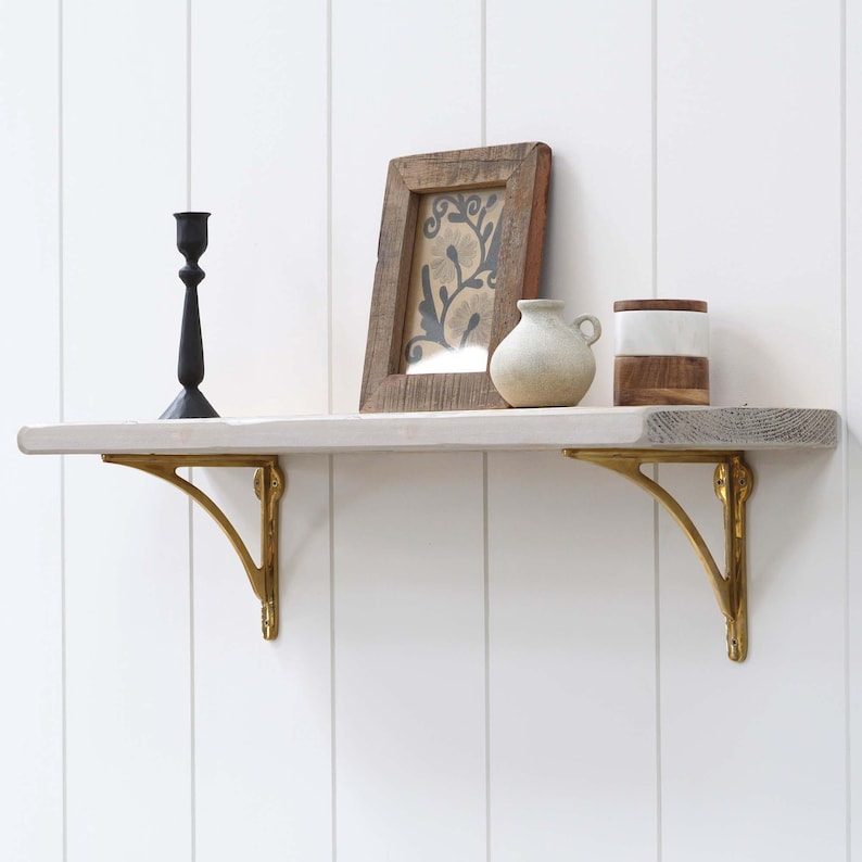 May include: A wooden shelf with gold-coloured brackets, displaying a black candlestick, a framed picture, a small vase, and a decorative container. The shelf is painted white and grey, mounted on a white panelled wall.