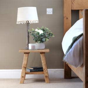 May include: A rustic wooden side table with a lamp, plant, and books. The lamp has a white shade with beaded trim. A white potted plant and a candle sit on the table. The table is next to a wooden bed frame.