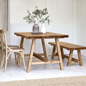 Könnte beinhalten: Ein rustikaler Esstisch aus Holz mit passender Bank und zwei Stühlen. Der Tisch ist etwa 152 cm lang und hat eine natürliche Holzoberfläche. Eine Vase mit Grün und ein weißer Krug stehen auf dem Tisch. Die Stühle haben geflochtene Sitze.