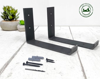 May include: Two black metal shelf brackets with mounting holes and screws. The brackets are on a white wooden surface with two gray plastic anchors and a drill bit.