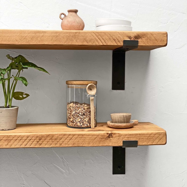 K&ouml;nnte beinhalten: Zwei Holzregale mit schwarzen Metallkonsolen. Das obere Regal hat einen kleinen Terrakotta-Topf und einen wei&szlig;en Teller. Das untere Regal hat ein Glas gef&uuml;llt mit Granola, einen Holzl&ouml;ffel und eine kleine Holzschale.