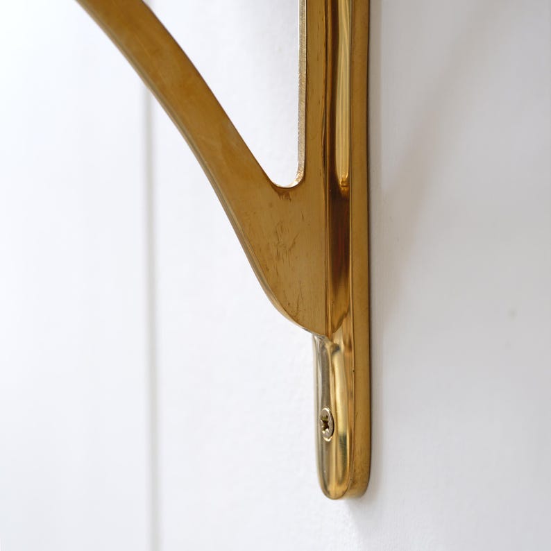 May include: A close-up of a gold-coloured metal shelf bracket. The bracket has a curved design and is mounted on a white wall. The bracket is made of a shiny, polished metal.