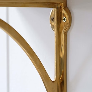 May include: Close-up of a gold-coloured shelf bracket. The bracket is mounted on a white wall and features a curved support arm. The bracket is made of metal and has a shiny finish. The bracket is attached to the wall with screws.