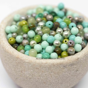 May include: A bowl filled with a variety of green and blue glass beads. The beads are round and have a smooth, polished finish.