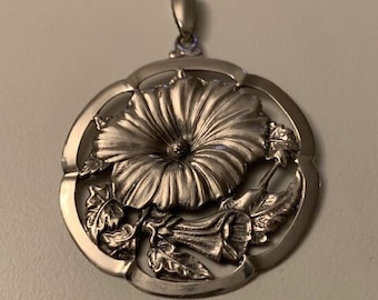 May include: A silver pendant featuring a detailed, three-dimensional design of a flower with leaves. The pendant is round and has a bail at the top for attaching to a chain.