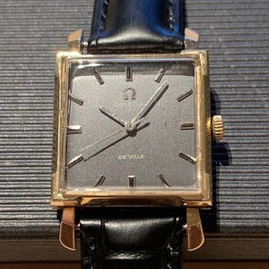 Könnte beinhalten: Eine goldfarbene Omega De Ville Armbanduhr mit einem schwarzen Lederband und einem quadratischen Zifferblatt. Das Zifferblatt hat einen silbernen Hintergrund mit schwarzen Stundenmarkierungen und Zeigern. Das Wort "De Ville" ist in Schwarz am unteren Rand des Zifferblatts aufgedruckt.