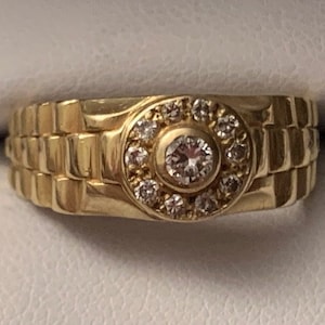 Pode incluir: Um anel de ouro com um grande diamante redondo cercado por diamantes menores. A banda do anel é de ouro texturizado.