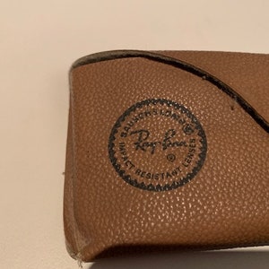 Peut inclure: Un &eacute;tui en cuir marron pour lunettes de soleil avec un logo Ray-Ban noir et le texte "Impact Resistant Lenses".