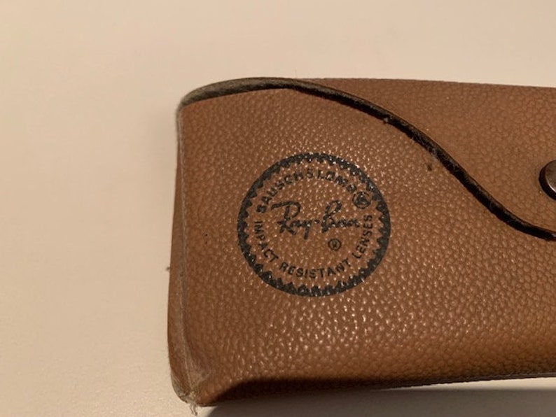 Peut inclure: Un &eacute;tui en cuir marron pour lunettes de soleil avec un logo Ray-Ban noir et le texte "Impact Resistant Lenses".