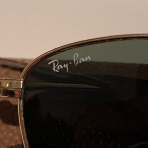 Peut inclure: Gros plan sur une paire de lunettes de soleil &agrave; monture dor&eacute;e avec des verres vert fonc&eacute;. Les lunettes portent l'inscription "Ray-Ban" sur les verres.