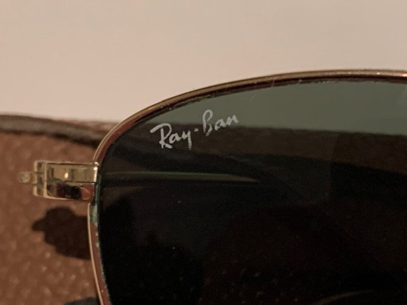 Peut inclure: Gros plan sur une paire de lunettes de soleil &agrave; monture dor&eacute;e avec des verres vert fonc&eacute;. Les lunettes portent l'inscription "Ray-Ban" sur les verres.
