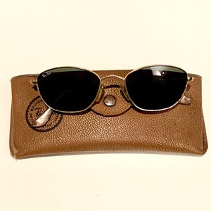 Peut inclure: Une paire de lunettes de soleil &agrave; monture dor&eacute;e avec des verres noirs dans un &eacute;tui en cuir marron. L'&eacute;tui porte le logo Ray-Ban en relief.