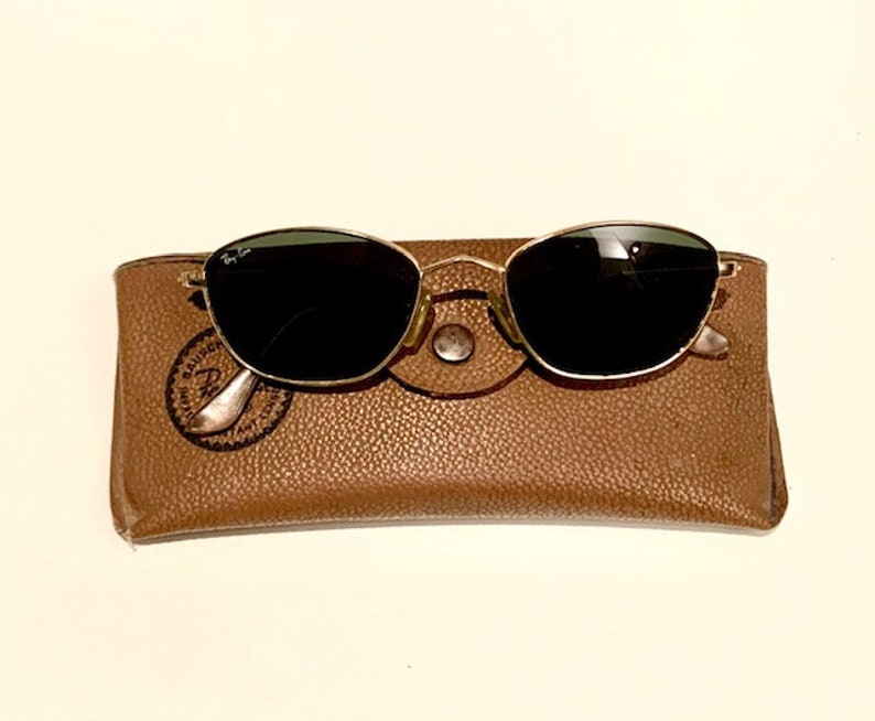 Peut inclure: Une paire de lunettes de soleil &agrave; monture dor&eacute;e avec des verres noirs dans un &eacute;tui en cuir marron. L'&eacute;tui porte le logo Ray-Ban en relief.