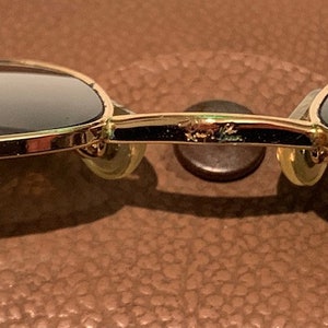 Peut inclure: Lunettes de soleil aviateur dor&eacute;es avec des verres fonc&eacute;s. Les lunettes ont une monture en m&eacute;tal et reposent sur une surface en cuir marron. Le nom de la marque "Ray-Ban" est visible sur le pont des lunettes.