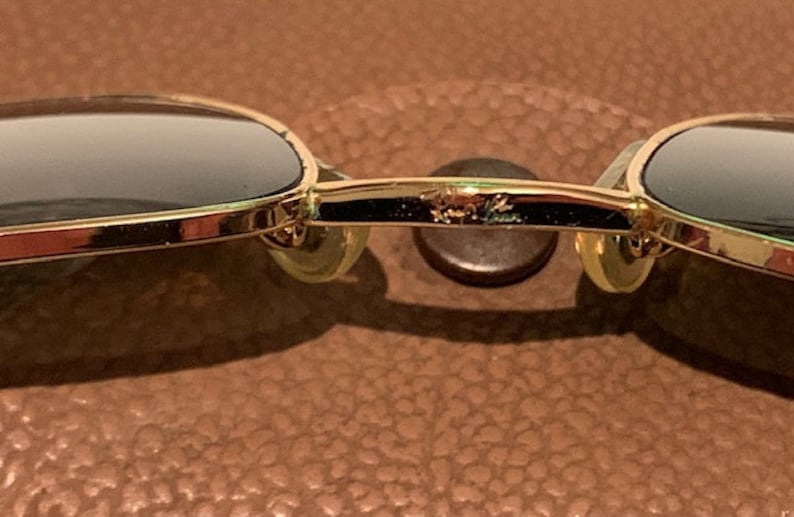 Peut inclure: Lunettes de soleil aviateur dor&eacute;es avec des verres fonc&eacute;s. Les lunettes ont une monture en m&eacute;tal et reposent sur une surface en cuir marron. Le nom de la marque "Ray-Ban" est visible sur le pont des lunettes.