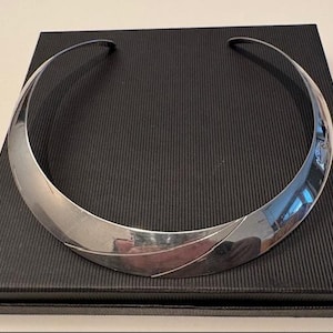 Könnte beinhalten: Ein poliertes silbernes Halsband mit geschwungenem Design, das auf einer schwarzen, strukturierten Schachtel liegt. Die Halskette hat eine glatte, reflektierende Oberfläche, und die Schachtel hat ein geripptes Muster.