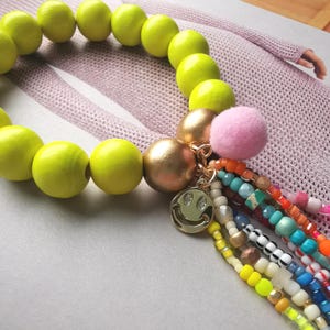 Könnte beinhalten: Ein lebendiges Armband mit großen, limettengrünen Holzperlen. Akzentuiert mit goldfarbenen Perlen, einem rosa Pompon und einem goldenen Smiley-Anhänger. Bunte Perlenquasten verleihen diesem einzigartigen Schmuckstück eine verspielte Note.