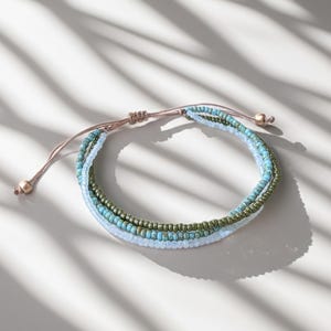 Könnte beinhalten: Ein mehrreihiges Armband mit drei Reihen kleiner Perlen in den Farben Hellblau, Türkis und metallischem Olivgrün. Das Armband wird mit einer dünnen, verstellbaren beigen Kordel befestigt, die an den Enden zwei kleine Metallperlen aufweist.