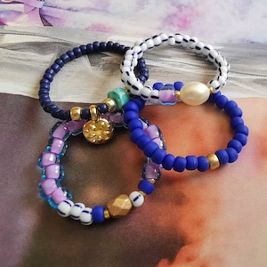 Könnte beinhalten: Vier Perlenarmbänder in Blau, Weiß und Lila mit einem goldenen Anhänger und einer weißen Perle. Die Armbänder sind übereinander gestapelt.