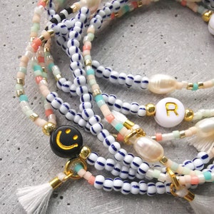 Könnte beinhalten: Mehrreihiges Perlenarmband mit weißen und blauen Perlen, rosa und türkisfarbenen Akzenten und goldenen Charms. Die Charms beinhalten einen Stern, eine Perle, ein Smiley und einen Buchstaben "R".