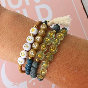 Op de afbeelding: Een set van vijf kralenarmbanden met gouden letters die de woorden "LOVE", "PEACE", "HOPE", "FAITH" en "JOY" spellen. De armbanden zijn gemaakt van kralen in verschillende kleuren, waaronder bruin, blauw en transparant.