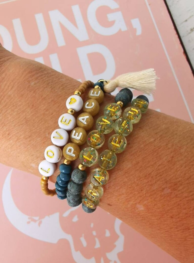 Op de afbeelding: Een set van vijf kralenarmbanden met gouden letters die de woorden "LOVE", "PEACE", "HOPE", "FAITH" en "JOY" spellen. De armbanden zijn gemaakt van kralen in verschillende kleuren, waaronder bruin, blauw en transparant.