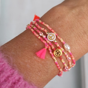 Könnte beinhalten: Vier übereinanderliegende Armbänder mit rosa, weißen und cremefarbenen Perlen. Die Armbänder haben einen Peace-Zeichen-Anhänger, einen Smiley-Anhänger und einen Perlenanhänger. Die Armbänder haben rosa Quasten.