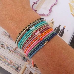 Könnte beinhalten: Ein Stapel bunter Perlenarmbänder am Handgelenk einer Person. Die Armbänder bestehen aus kleinen, runden Perlen in verschiedenen Blautönen, Grün, Rosa, Orange und Braun. Einige der Perlen sind golden.
