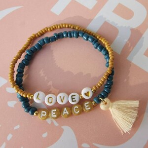Op de afbeelding: Een dubbele rij kralen armband met een gouden hart bedel en een witte kwast. De kralen zijn blauw en goud, en de armband spelt "LOVE PEACE" in witte letters.