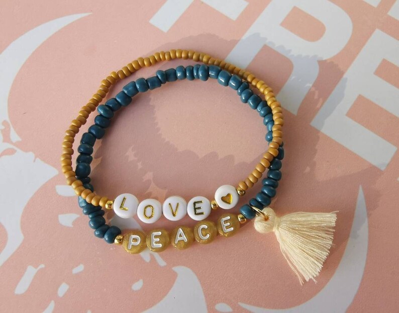 Op de afbeelding: Een dubbele rij kralen armband met een gouden hart bedel en een witte kwast. De kralen zijn blauw en goud, en de armband spelt "LOVE PEACE" in witte letters.