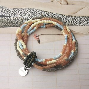 Könnte beinhalten: Mehrreihiges Perlenarmband mit einem silbernen Anhänger, auf dem "Calmer_and_co" steht. Das Armband besteht aus braunen, beigefarbenen, hellblauen und grauen Perlen.