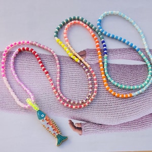 Könnte beinhalten: Drei farbenfrohe Perlenketten. Eine Halskette hat einen Fischanhänger mit orangefarbenen, blauen und weißen Details. Die anderen beiden Halsketten bestehen aus kleinen, farbigen Perlen in Rosa-, Grün-, Blau- und Orangetönen.