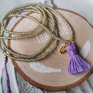 Könnte beinhalten: Eine hellgrüne Perlenkette mit einem violetten Quastenanhänger. Die Kette ist um eine Holzscheibe gewickelt, auf der die Wörter "the" und "little" stehen.
