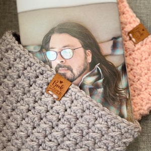 May include: A gray crocheted book cover with a brown leather tag that says "Book Lover" on it. The cover is holding a magazine with a man wearing glasses on the cover.