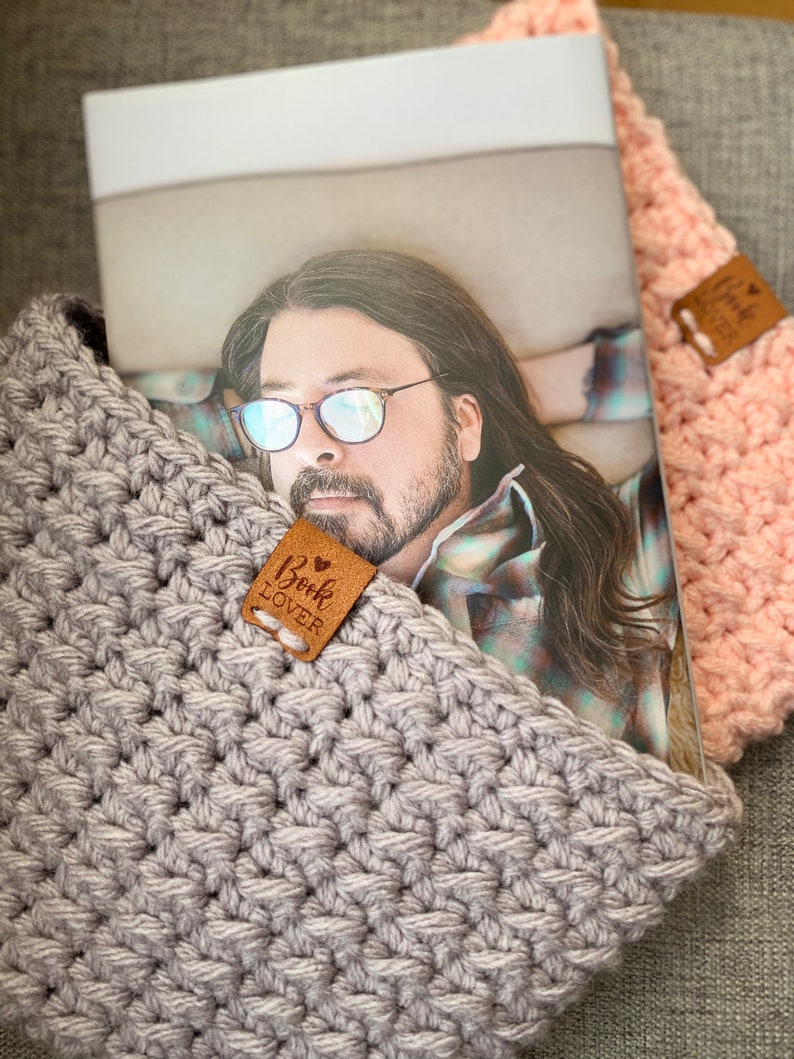 May include: A gray crocheted book cover with a brown leather tag that says "Book Lover" on it. The cover is holding a magazine with a man wearing glasses on the cover.