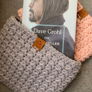 May include: A gray crocheted book sleeve with a brown leather bookmark that says "Book Lover" is holding a book titled "Dave Grohl The Storyteller".