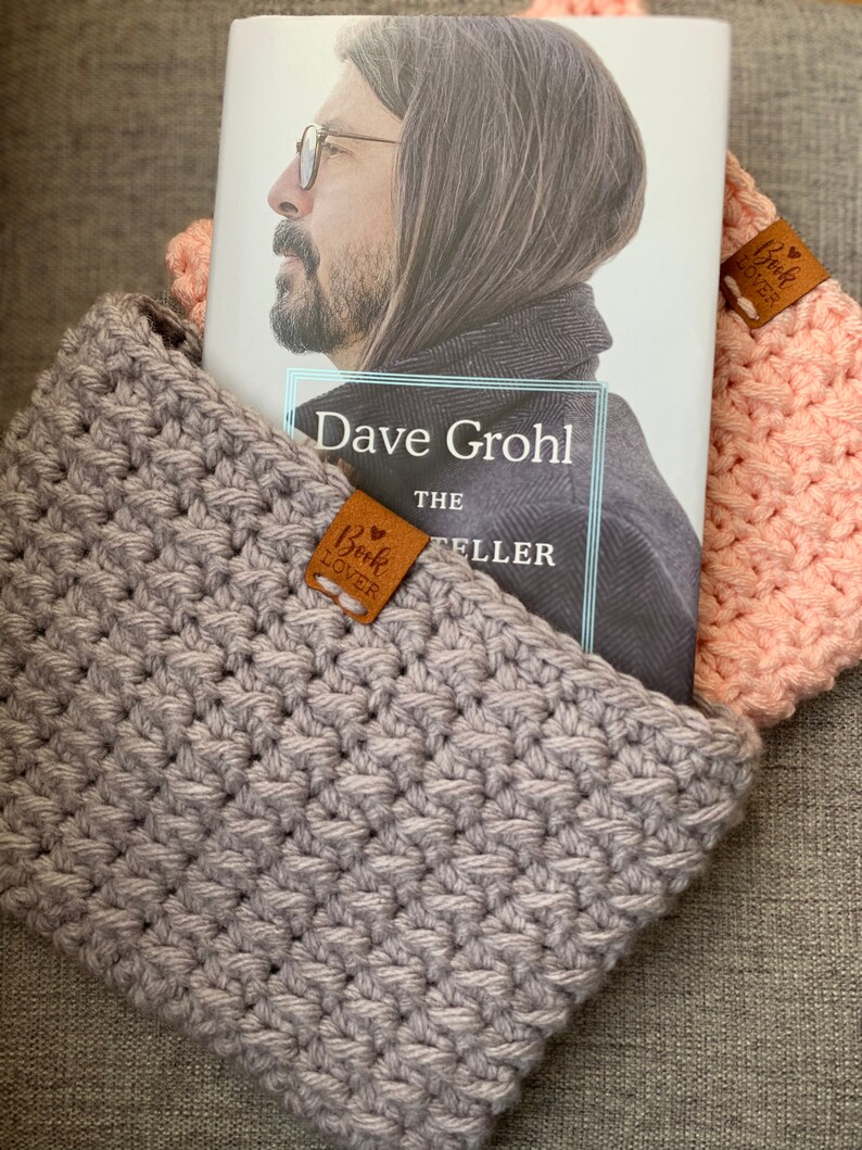 May include: A gray crocheted book sleeve with a brown leather bookmark that says "Book Lover" is holding a book titled "Dave Grohl The Storyteller".