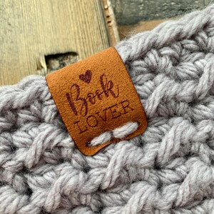May include: A brown leather tag with the text "Book Lover" and a small heart stitched on it. The tag is attached to a gray crocheted item.