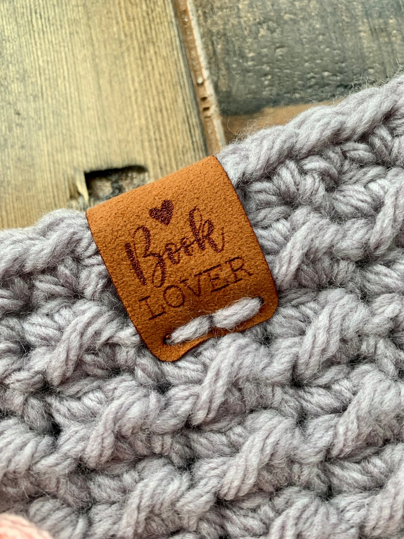 May include: A brown leather tag with the text "Book Lover" and a small heart stitched on it. The tag is attached to a gray crocheted item.