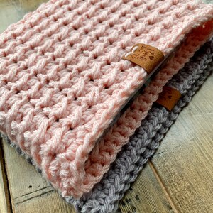 May include: Two crocheted scarves, one pink and one gray, are folded together on a wooden surface. The pink scarf has a brown leather tag with the text "Forever Grateful" on it.