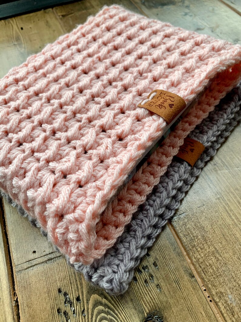 May include: Two crocheted scarves, one pink and one gray, are folded together on a wooden surface. The pink scarf has a brown leather tag with the text "Forever Grateful" on it.