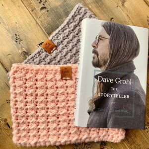 May include: Two crocheted neck warmers in pink and gray with leather tags that say "Book Lover". A book titled "Dave Grohl: The Storyteller" is on top of the warmers.