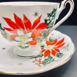 May include: A white teacup and saucer set with a Christmas poinsettia design. The cup and saucer feature red poinsettia flowers, green holly leaves, and gold accents.