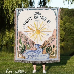 May include: A woven tapestry throw blanket with a light blue background and a colorful illustration of a sun, a river, and plants. The text on the blanket reads "The Light Shines in the Darkness but the Darkness has not overcome it."